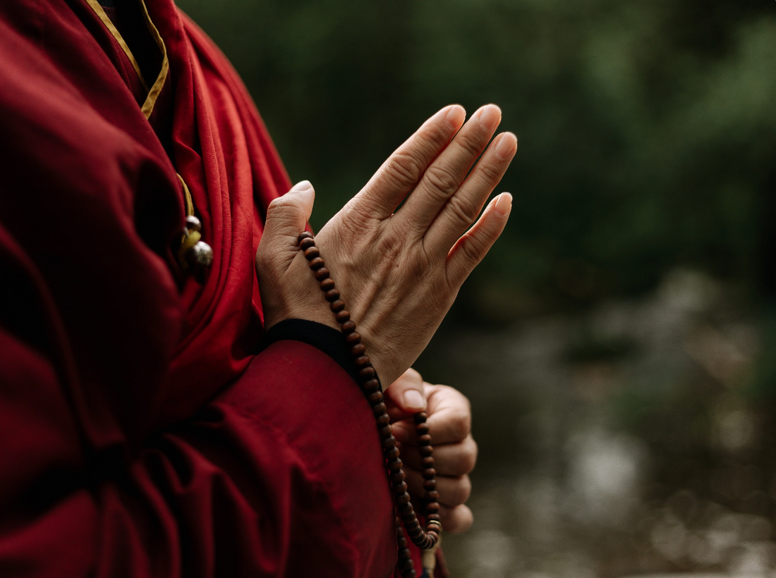  Ein Foto, das einen tibetischen Mönch in roter Robe zeigt, der eine Gebetskette (Mala) in der Hand hält.